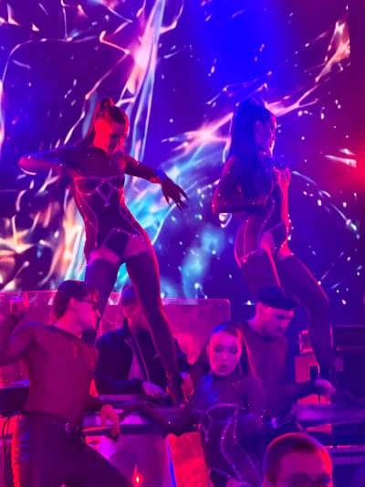 This image depicts a dimly lit indoor stage performance, possibly in Tsilkani, Georgia. The background is a vibrant, abstract light show with swirling patterns in shades of purple, blue, and pink. Two female performers in sparkling, dark, form-fitting outfits are center stage, captured in mid-dance. The performer on the left has her hair in a high ponytail and her arms extended. The performer on the right is also in motion. In the foreground, three individuals are visible. A male performer wearing a sheer, sparkly top and black leather pants is on the left, his left arm raised. Another male figure, wearing a dark beret and a dark long-sleeved shirt, is behind him and to the right, facing towards the performers. A third person, whose face is mostly obscured and out of focus, is in the lower right corner, wearing glasses. The stage appears to have some equipment, possibly musical instruments or DJ decks, in front of the performers. The overall atmosphere is energetic and theatrical, with dramatic lighting.
