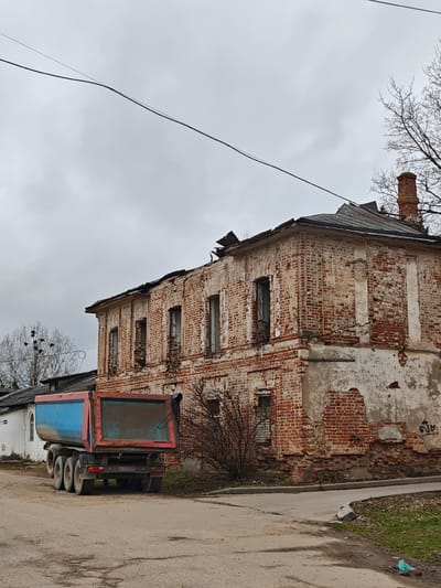 The image depicts a dilapidated two-story brick building with missing windows and crumbling facade, located in посёлок совхоза "Буденновец", Russia. A dump truck with a blue and red bed is parked on a dirt road in front of the building. The sky is overcast. A few bare trees are visible. A white fence is partially seen on the left. A power line stretches across the top portion of the image. Graffiti is visible on the lower portion of the building's right side. The building appears abandoned and in disrepair.
