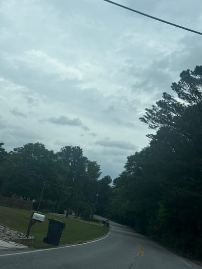 The image shows an overcast sky above a residential street in Mountain Park, United States of America. Power lines are visible against the sky. The street curves, bordered by trees and green lawns. A mailbox with a visible address of "1866" stands near the roadside. The road surface appears wet, possibly from rain.
