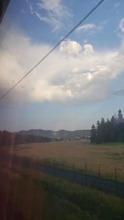 This is a landscape photograph taken from inside a vehicle, likely a train, as indicated by the reflection on the left side of the image and the blurred foreground. The scene depicts a rural area with a vast, open field in the mid-ground, appearing golden or tan, suggesting it might be a harvested crop or dry grass. A dark, unpaved road runs horizontally across the middle ground, bordered by a chain-link fence with visible posts.
In the background, rolling hills covered in dark green forests are visible. A small cluster of buildings and structures, possibly a village or isolated houses, can be seen nestled among the trees on the far right side of the background. A line of evergreen trees stands on the far right edge of the image, some of them tall and dark.
The sky is a prominent feature, filled with a mix of bright blue patches and large, fluffy white clouds. Towards the left side of the sky, there are darker, more diffused clouds, possibly indicating approaching or receding rain, with a faint hint of a rainbow or atmospheric effect. Two thin, dark wires or cables stretch diagonally across the upper portion of the image, from the top left corner towards the middle right.
The location is identified as Lučivná, Slovakia. The overall impression is one of a serene, rural landscape under a dynamic sky.