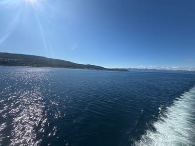 The image depicts a bright, sunny day at sea, likely near Bognes, Norway. The scene is dominated by a vast expanse of deep blue water, shimmering with reflected sunlight. The sun itself, a brilliant white orb, is prominently featured in the upper left corner, casting strong rays that create a lens flare effect across the clear blue sky.
In the background, a green, hilly coastline stretches across the horizon. The hills appear to be covered in vegetation and dotted with small buildings, suggesting a settled area. Further in the distance, hazy mountains can be seen, some with snow-capped peaks, indicating a mountainous region. The water in the foreground is marked by a distinct white wake trailing from the right side of the frame, suggesting the perspective is from a moving vessel, possibly a ferry. The wake is foamy and turbulent, contrasting with the calm surface of the ocean further out.
The overall atmosphere is one of tranquility and natural beauty, amplified by the clear, sunny weather. The time of day appears to be daytime, given the strong sunlight and clear skies. There is no visible text in the image.