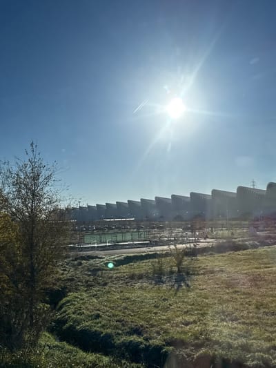 The image captures an exterior scene in Reggio nell'Emilia, Italy, focusing on the Reggio Emilia AV Mediopadana high-speed rail station. The station, positioned in the background, is characterized by its distinctive, elongated, white or light-grey undulating canopy segments, resembling a series of waves or sawteeth. In the midground, a large, flat area is visible, containing what appears to be a green-surfaced court, potentially for tennis or padel, adjacent to a parking lot filled with numerous parked vehicles. Sections of bare earth and minor construction or landscaping are also present. The foreground consists of a grassy field, displaying a frosted or dewy appearance, indicating cool temperatures. A small, dry ditch or stream bed traverses the lower left portion of this field, near a leafless deciduous tree. The sky is clear and blue, with a prominent, low-angle sun in the upper right quadrant generating substantial lens flare and a visible aircraft contrail to its left. No individuals are discernible within the frame.