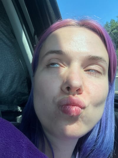A close-up, slightly angled selfie of a fair-skinned woman with purple hair. Her eyes are partially closed, and her lips are pursed. The background shows a glimpse of blue sky and the interior of a vehicle. The exact location in the city of Pourrières, France, is indeterminable from the image.