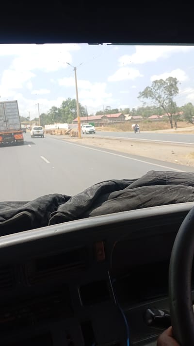 View from a vehicle driving on a paved road with other vehicles present. A large truck is in the left lane, and two white cars are visible ahead. There's roadside construction in progress. A motorcyclist travels on the right. The dashboard and steering wheel of the vehicle are visible, with dark cloth covering parts of the dash. Utility poles line the roadside, with buildings and trees in the background. The sky is light blue with some clouds.