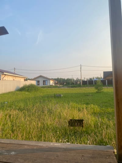 The image depicts a residential construction area in Raskaty, Russia, under a clear sky with wispy clouds. In the foreground, a wooden surface, possibly a table or deck, is visible. Beyond this, a large expanse of tall, green grass covers the ground. Several houses are in various stages of construction or completion. To the left, a house with a brown roof and white brick walls is partially obscured by a pale fence. Further back and to the right, a single-story house with a light-colored facade and dark-framed windows is prominent. Other structures, including fences and trees, are scattered throughout the landscape. Power lines run across the background. The scene appears to be captured during daylight, likely late afternoon, given the warm lighting.