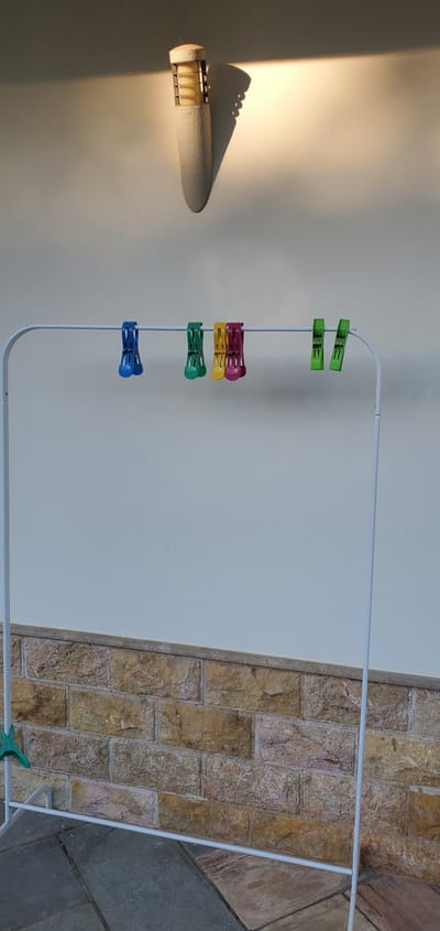 The media file shows a clothesline with several colorful clothespins attached to it. The clothesline is white and appears to be made of metal. There are green, pink, yellow, and blue clothespins. The background is a white wall, and on the right side of the image, there is a textured wall made of stones. In the upper left corner, a light fixture casts a shadow on the white wall. The lighting suggests it is daytime, possibly late afternoon due to the warm glow and long shadow. The scene appears to be outdoors, possibly in a backyard or patio area. The overall impression is one of domesticity and everyday life. No people are visible in the image.
