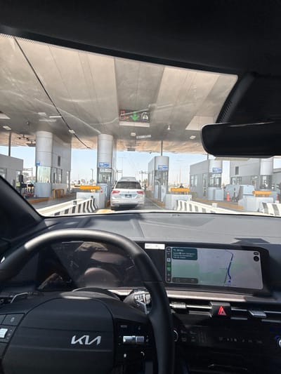An interior view of a Kia vehicle at a toll plaza, showing multiple toll booths. A white vehicle waits ahead. The dashboard displays a navigation screen with a route. The steering wheel with the Kia logo is visible. The car is at a standstill.