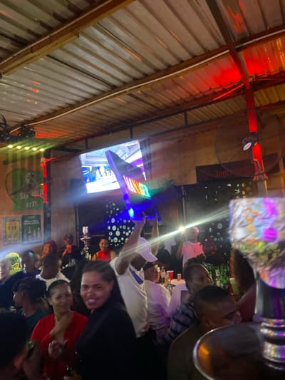 The image shows a crowded bar in Dodoma, Tanzania, located at coordinates -6.157 latitude and 35.724 longitude. The bar is dimly lit, with the ceiling covered in corrugated metal panels. A man in a white shirt and a white hat holds a colorful sign with the word "WINNER" in bold letters. Several other people are seated at tables, many of whom appear to be enjoying beverages. There are also numerous bottles of beer and other drinks visible throughout the scene. There is a large television screen in the background, but the image is too blurry to determine what is being displayed on it. Overall, the image depicts a lively, celebratory atmosphere.