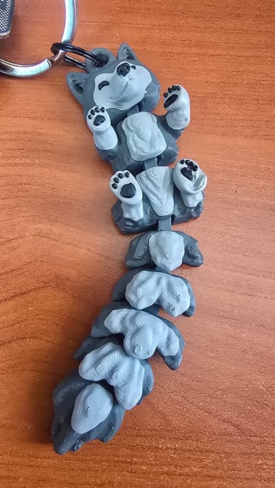 A gray and white wolf keychain is lying on a wooden surface. The wolf is depicted in a playful pose, with its legs and paws raised in the air. It is connected to a silver keyring. The wooden surface is a light brown color with visible grain. The lighting suggests an indoor environment, possibly on a table or desk. There is no indication of the city of Krasnopol'ye, Russia, in the image.