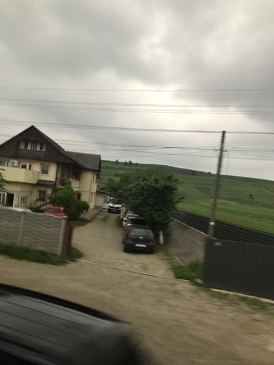The image displays a daytime view from Cristești, Romania, under an overcast sky. The foreground reveals the top of a dark vehicle dashboard or window frame, suggesting the photograph was taken from inside a moving car.
In the midground, a two-story house with a brown gabled roof and light-yellow walls is visible on the left. The house features multiple windows, an upper-floor balcony with railings, and an external air conditioning unit mounted on the upper wall. A small red car is partially visible near the house, partially obscured by green foliage.
A dirt or gravel driveway extends from the foreground towards the background, passing the house. Several vehicles are parked along this driveway, including a dark blue hatchback on the right, and a white van further down the drive. Green trees and bushes are interspersed along the driveway and around the house. A low, grey concrete fence borders the left side of the driveway near the house.
The background consists of rolling green hills, some with visible patches of darker green vegetation, extending to the horizon under the cloudy sky. A wooden utility pole with multiple parallel power lines runs vertically on the right side of the image, extending into the sky. A dark, corrugated fence, possibly metal, runs horizontally along the right edge, separating the driveway area from the distant green landscape. No individuals are visible in the scene.