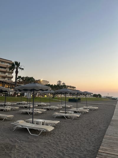 The image shows a beach in Rhodes, Greece, at dusk. The sky is a clear gradient of pale blue to soft orange near the horizon. In the foreground, rows of empty, light-colored lounge chairs with matching small tables are set up on a dark sand beach. Each chair is equipped with a beige cushion. Behind the lounge chairs, several large, dark grey umbrellas are erected, some open and providing shade. Further back, a strip of green grass separates the beach from a modern hotel building with multiple floors, balconies, and palm trees. The hotel's facade is lit by artificial lights. The right side of the image features a wooden boardwalk running parallel to the beach. The overall scene conveys a sense of tranquility and an invitation to relaxation.