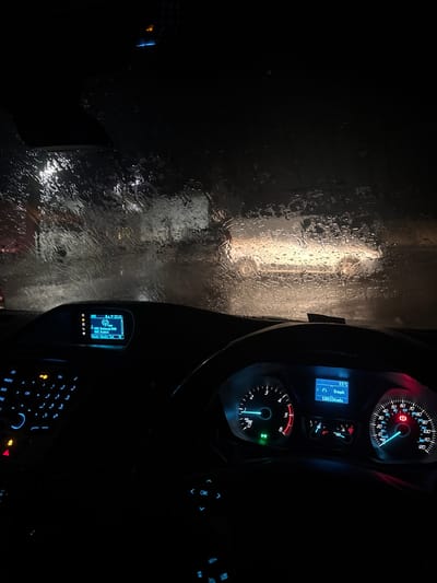The image depicts the interior of a stationary vehicle at night, viewed through a rain-streaked windshield in Sigglesthorne, United Kingdom. The dashboard is illuminated, showing various controls and displays. The central infotainment screen displays "BBC Radio 4" and "BBC Digital DAB" with the time "22:11". To the right, the instrument cluster's digital display indicates an ambient temperature of "15°C", a speed of "0 mph", and an odometer reading of "100334 mils". Both the tachometer and speedometer needles are at their lowest positions, consistent with a parked vehicle. Illuminated warning lights are visible on the instrument cluster. Outside the vehicle, the diffused bright headlights of at least one other vehicle are visible directly ahead through the heavily wetted windshield, suggesting traffic or a developed area. No individuals are visible.