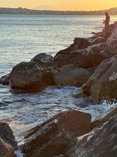 An elevated, medium shot captures a man fishing on a rocky shoreline in Alimos, Greece, during sunset. The man, silhouetted against the golden sky, stands on a cluster of large, dark rocks, holding a fishing rod. The water is calm with gentle waves lapping against the rocks, creating white foam. In the background, a hazy city skyline with buildings and a distant mountain are visible. The light casts a warm glow over the scene.