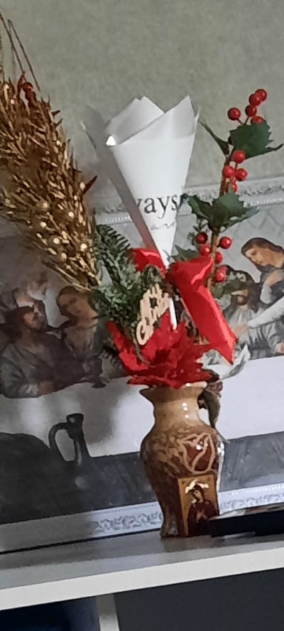 The image shows a close-up of a festive decoration. The main subject is a wooden reindeer figure, likely part of a larger holiday display. The reindeer is depicted from the side, with its head and part of its body visible. It appears to be made of dark wood with intricate, lighter-colored carvings or inlays on its surface, including what looks like a golden embellishment on its forehead.
Attached to the reindeer's neck is a decorative arrangement featuring faux holly with red berries and green leaves, along with a bright red ribbon tied into a bow. There's also a fluffy, red, flower-like accent and a small, wooden ornament shaped like a star or snowflake with the word "Merry" inscribed on it. Below this, a sprig of green pine needles and a bundle of what appears to be dried reeds or straw, possibly adorned with tiny white lights, complete the festive ensemble. A white cone-shaped object, possibly part of the packaging or another decorative element, is also visible to the left, with the partial word "ways" printed on it.
The background is blurred, but it seems to be a patterned surface, possibly wallpaper or fabric, with muted colors and what might be an illustration of animals. The lighting suggests an indoor setting, and the overall impression is one of a holiday or seasonal decoration. There are no people visible, and no specific time of day or weather conditions can be inferred. The location context provided is Bocani, Moldova, suggesting this decoration might be found in a home or public space there.