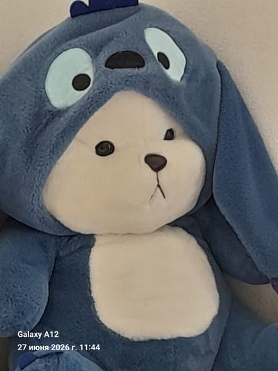 A close-up, eye-level, medium shot captures a blue plush toy with a white belly. The toy is wearing a blue hood with two large, light blue eyes, each with a black pupil. Above the eyes, on the hood, are two small black shapes that resemble eyebrows or part of the toy's character design. The toy has a white snout with a small black nose and a thin, dark line representing its mouth, which is slightly curved upwards. The background is a plain, light-colored surface. In the bottom left corner, text reads "Galaxy A12" and "27 июня 2026 г. 11:44," indicating the device and date/time of the photograph. The toy appears to be placed indoors.