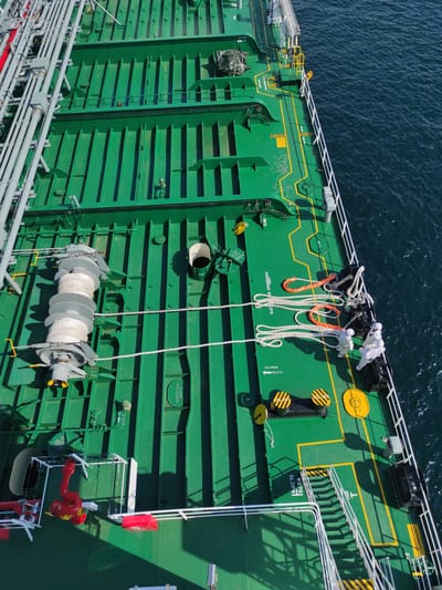 This elevated aerial view captures a brightly lit ship deck, likely a tanker, in Kunak, Malaysia, under clear sunny skies. The deck is predominantly green, marked with yellow lines indicating pathways and areas. Two individuals, clad in white protective suits, are seen on the upper deck, seemingly engaged in handling or preparing large white ropes, some of which are covered in orange sheathing. These ropes are coiled and laid out near black bollards with yellow safety markings.

A large winch or capstan with a thick, white rope coiled around it is visible on the lower deck, alongside numerous grey and red industrial pipes running horizontally across the vessel's structure. The ship's side features prominent horizontal ribbing. Various hatches, including yellow circular ones labeled "ULL HOLE SLIPS," are scattered across the deck. A white funnel-like structure protrudes from the side, and a red fire hose or emergency equipment is mounted on a white platform.

Visible text on the deck includes "PLOT ACCESS POINT KEEP CLEAR," "WB7S," "SLOPS," "COGS RSDO ROOPS" (partially obscured), "ULL HOLE SLIPS," and "SLOPS NOLADDER." Another sign reads "DO NOT OPERATE." The deep blue, rippled ocean surrounds the vessel, reflecting the bright daylight. The scene suggests active maritime operations, possibly involving mooring or cargo handling, in a busy port or offshore location.