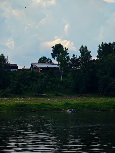 A wide shot captures a tranquil scene of a body of water, likely a river, in Ust-Kem', Russia, under a cloudy sky. The water's surface is rippled, reflecting the muted light. On the far bank, a grassy slope is densely wooded with various trees, including birch and pine. Several wooden structures, resembling houses or outbuildings, are visible nestled amongst the trees on the elevated terrain. The architecture of the buildings appears rustic, with visible wooden siding and pitched roofs. In the foreground on the water, a small motorboat is moored near the shore, resting on the grass by the water's edge. The overall atmosphere is one of rural serenity.