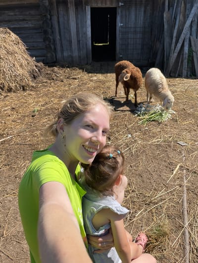 A bright, sunny day finds a woman in a lime green t-shirt taking a selfie, holding a young child on her hip. The woman is smiling widely, with blonde hair pulled back and a nose ring. The child, seen from behind, has dark hair tied with light blue scrunchies. In the background, two sheep are grazing on a pile of daisies in a rustic farmyard, near a weathered wooden barn. Hay is scattered on the ground, and a large haystack is visible to the left. The scene appears to be set in the village of Syam-Mozhga, Russia.