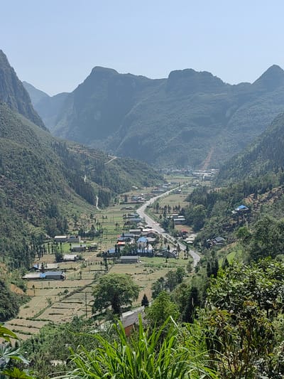 The image depicts a broad valley in the Phố Bảng Commune, Vietnam, nestled amongst rolling, verdant mountains under a clear sky. A winding road cuts through the center of the valley, flanked by rows of houses with assorted colored roofs and agricultural fields arranged in stepped terraces. Trees are scattered throughout the scene. The foreground is dominated by dense vegetation.