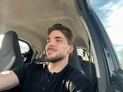 The image shows a man with brown hair and a beard wearing a black shirt with buttons, seated inside a vehicle. The interior of the vehicle includes a beige roof, grey and black seats, and a black frame around the window. The sky is visible through the window. The man is wearing a seatbelt.
There is no definitive evidence to confirm the location as Volketswil, Switzerland, based solely on the image.