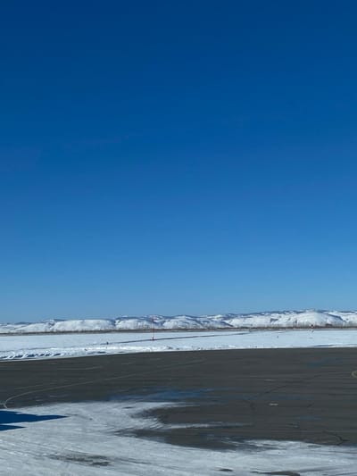 The image, when properly oriented (rotated 90 degrees counter-clockwise), displays a wide-angle landscape primarily composed of a vast blue sky, distant snow-capped mountains, and a flat, dark foreground.
The upper portion of the image is dominated by a clear, deep blue sky, which fades slightly to a lighter hue closer to the horizon. There are no clouds visible, indicating a bright and sunny day.
Stretching across the midground, just below the sky, is a long, undulating range of snow-capped mountains or large hills. These mountains appear distant and rugged, their white peaks and slopes contrasting sharply with the deep blue sky.
The lower half of the image features a large, flat, dark-colored ground surface, resembling an asphalt runway, a vast paved area, or a large parking lot. This surface shows visible cracks and is partially covered with patches of snow and ice, suggesting cold or wintery conditions. Long, distinct dark shadows are cast across this paved area, extending from the right side of the frame towards the left, indicating bright sunlight likely from a low angle, such as morning or late afternoon. The shapes of these shadows suggest they might be cast by structures or objects just outside the frame.
Between the dark foreground and the distant mountains, there is a narrower strip of lighter, snow-covered ground, which could be a cleared path, road, or simply a flatter expanse of snow. Within this strip, two slender poles, each topped with a small red flag or marker, are sparsely visible, adding minor points of interest to the otherwise desolate scene.
The overall impression is one of a vast, open, and cold environment with clear weather. There are no people, animals, or significant man-made structures (apart from the paved surface and small poles) visible in the frame, conveying a sense of isolation and natural expanse. No text is visible in the image.