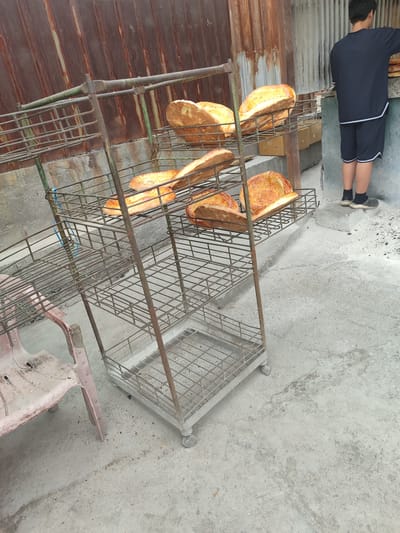 In a city setting of Tokmok, Kyrgyzstan, a young person stands near a metal rack displaying several loaves of bread. The bread appears golden brown and crusty. The rack is made of a gray metal with multiple shelves, and some of the shelves have bread on them. A dusty pink plastic chair is visible on the left side of the image, and to the right, the young person, wearing a dark t-shirt and shorts, is facing away from the camera. The background consists of a weathered, corrugated metal wall and a concrete ground.