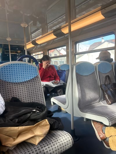 The image depicts the interior of a public transport vehicle, likely a train or tram, located in Villemomble, France. Three individuals are visible within the cabin. In the mid-ground, a woman with dark hair, wearing a red long-sleeved top and dark pants, is seated. She holds a smartphone in her right hand and has her left hand pressed against her forehead. A white earbud is visible in her right ear. A grey blanket or garment rests on her lap, and a blue tote bag occupies the seat next to her. Further back and to the right, the rear view of a person wearing a brown baseball cap and dark clothing is visible; a dark brown leather shoulder bag hangs on the seat directly in front of them. In the immediate foreground, the lower legs and feet of a third passenger are shown, dressed in tan pants, striped socks, and brown shoes. A black garment and a crumpled brown paper bag are placed on the seat in front of this foreground passenger. The vehicle's interior features patterned grey and blue seating, overhead fluorescent lighting, and vertical support poles. Through the windows, residential buildings with pitched roofs and a clear blue sky are visible, indicating an urban or suburban setting.