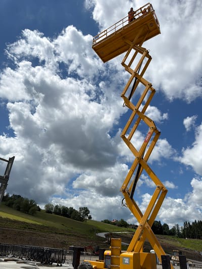 A yellow scissor lift, raised high into a partly cloudy blue sky, is the primary subject. Two individuals are visible in the lift's platform at the top. The scissor lift's complex geometric structure is prominent, extending upwards from a base unit on the ground. The surrounding environment appears to be a construction site or outdoor event setup. In the background, a green, hilly landscape with trees and some buildings can be seen. Fencing and other construction equipment are present on the ground level. The image appears to be taken on a bright, possibly sunny day, with dramatic cloud formations. The specific location of Janowice, Poland is not discernible from the visual information within the image itself.