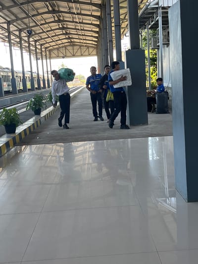A group of four individuals are on a train station platform in Desa Tambakreja, Indonesia. The platform is covered by a metal roof supported by large pillars. A train is visible on the tracks to the left. The first individual, wearing a white shirt and black pants, is carrying a large, green, round bag over his left shoulder. Behind him, a second individual in a blue uniform shirt and black pants is walking. To the right of the second individual, a third person in a blue shirt and black pants is carrying a white, rectangular package. A fourth individual, also in a blue shirt and black pants, is further to the right, near a large black trash bag. The ground of the platform is tiled and reflective. Several potted plants are positioned along the edge of the platform. Sunlight is casting shadows and illuminating the scene.