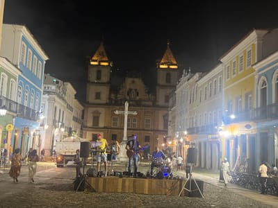 The image depicts a nighttime scene in the Largo do Cruzeiro de São Francisco, located within the Pelourinho district of Salvador, Brazil. A live music performance is taking place on a raised wooden stage situated on the cobblestone square. Three male musicians are actively performing: a keyboardist wearing a yellow shirt and a red cap, a guitarist in dark attire, and a drummer dressed in a blue shirt. Musical instruments and equipment, including a keyboard, guitars, a drum kit, microphones, and speakers on stands, are arranged on and around the stage, illuminated by stage lights.
Behind the performing band, a large white stone cross stands centrally in the square. Dominating the background is the illuminated Igreja e Convento de São Francisco (Church and Convent of Saint Francis), distinguished by its ornate façade and two prominent bell towers. Historic colonial-style buildings, featuring multiple stories, balconies, and white-framed windows, line both sides of the square. These buildings are painted in various bright colors, including blue, green, yellow, and white.
Several pedestrians are present on the cobblestone street; two women are visible walking on the left side, and other individuals are scattered further back, some seated at outdoor tables on the right. A light-colored truck with a raised bed is positioned on the left side of the square, behind the pedestrians. The entire scene is artificially illuminated by streetlights, shop lights, and stage lights under a dark night sky.