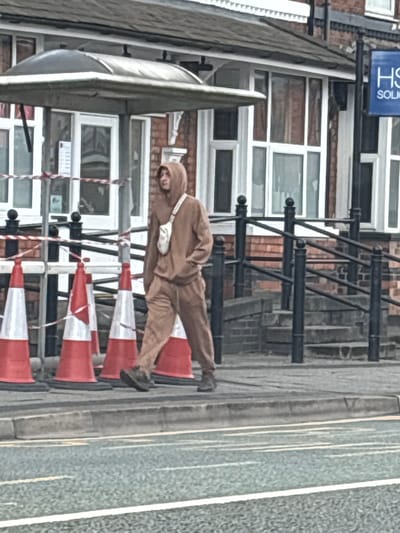 A man wearing a brown hooded jumpsuit and a white shoulder bag is lying horizontally across a row of orange and white traffic cones on a pavement. The pavement is adjacent to a road with white line markings. Behind the cones, there is a building with multiple windows, a black metal railing, and a blue sign that says "HS" in white lettering. The location is Crewe, United Kingdom.