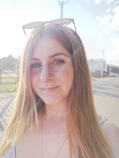 A young woman with blonde hair and freckles is shown in a close-up selfie, taken outdoors during the day, with the sun in the background creating lens flare. She is wearing sunglasses on top of her head and a necklace. The background shows a street with some buildings and a fence, suggesting an urban or suburban environment. There are clouds in the sky, indicating a partly cloudy day. The location is identified as Ivanovskoe, Russia.
