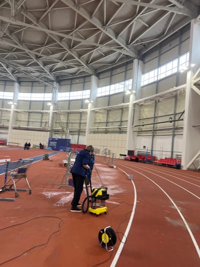 This image presents an interior view of an indoor athletic facility in Tashevka, Russia. The scene is dominated by a red track surface with white lane markings, encompassing a large, well-lit space. The ceiling consists of a complex network of light-colored metal trusses supporting a corrugated roof. The upper sections of the walls feature multiple rectangular windows allowing ambient light, supplemented by wall-mounted artificial lighting fixtures.

In the foreground, a single individual, a female wearing a dark blue short-sleeved top, dark trousers, and light-colored athletic shoes, is actively engaged in cleaning the track. She is wearing blue gloves and operating a yellow and black wet/dry vacuum cleaner with a metal drum, using a long hose attachment to suction white, foamy residue from the track surface. A black power cord extends from the vacuum, connecting to a black cable reel with yellow accents positioned on the track in the lower right. To the left of the cleaning operation, a silver-colored shopping cart is visible on the track, near what appears to be a hurdle.

The midground and background include elements characteristic of an athletic arena. Several metal barriers and hurdles are visible along the track. In the far background to the left, a blue mat area is present, with several indistinct figures seated or standing nearby. Along the right side of the track, red tiered spectator seating is visible. A tall, light-colored metal structure, possibly scaffolding or a support column, stands in the left midground. Another small yellow and black cleaning machine is situated further down the track on the left. The scene depicts an ongoing maintenance activity within the sports complex.