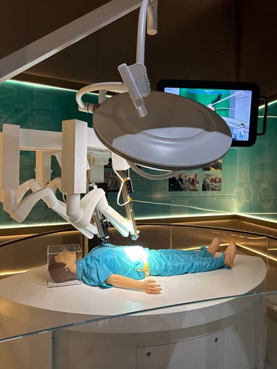 The image depicts a medical technology exhibition display located in Guangming District, China. A lifelike mannequin, dressed in light blue surgical scrubs and tan shoes, lies supine on a large, elevated, curved white platform. A multi-armed white robotic surgical system, similar to a Da Vinci system, is positioned directly above the mannequin's abdomen, with one metallic instrument arm extended as if engaged in a procedure. A prominent circular white and gray surgical light fixture is suspended overhead, casting bright illumination onto the simulated surgical site. To the right, a black rectangular monitor displays a green-toned graphical interface, likely illustrating an internal view or simulation. The mannequin's head is covered by a clear plastic box, and a yellow strap is secured around its waist. The background consists of a teal-green wall adorned with horizontal strip lighting, informational text panels, photographic images, and a stylized white electrocardiogram waveform graphic. A dark brown ceiling is visible, and a clear glass barrier runs across the immediate foreground, separating the display from the viewer.