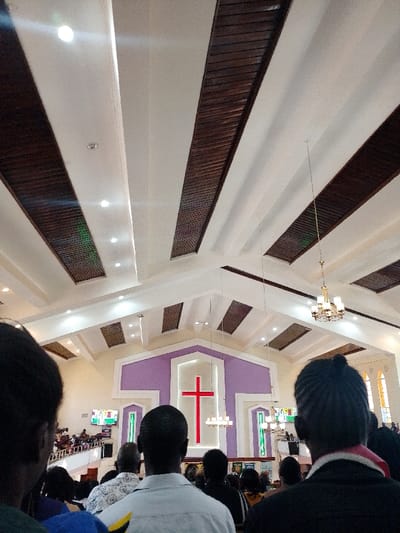 The image displays the interior of a church located in Karuri ward, Kenya, viewed from the rear towards the sanctuary. A prominent red cross is centrally positioned on the front wall, set against a multi-layered purple and white geometric backdrop. The high, vaulted ceiling is white, featuring parallel dark wood panels integrated with numerous recessed circular lights, and two brass-toned chandeliers are suspended. A large congregation of individuals is visible, primarily from behind, occupying the main floor and an elevated balcony section on the left. Two flat-screen monitors, displaying visual content, are mounted on the side walls near the front. Arched windows on the right wall permit natural light into the space. Individuals are oriented towards the front of the room.
