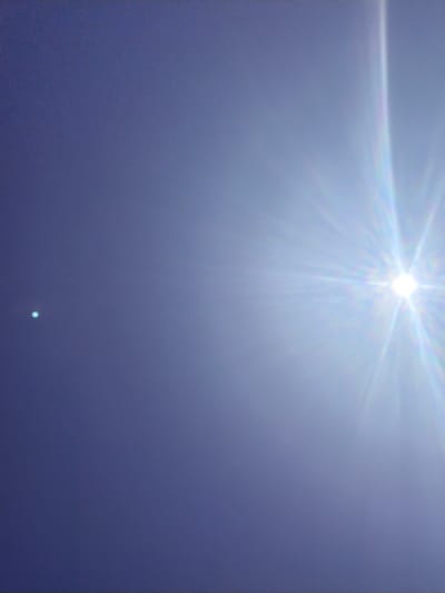 The image shows a clear blue sky with the sun prominently visible on the right side, emitting bright rays. A small, bright blue-green dot is visible in the left portion of the sky. The image appears to be taken outdoors in an open area, likely during daylight hours, possibly in the city of Povarovka, Russia, given the context of the prompt, though this cannot be definitively confirmed from the visual data alone. There are no people or man-made structures clearly identifiable in the frame. The overall scene depicts a sunny day with a celestial object in the sky.