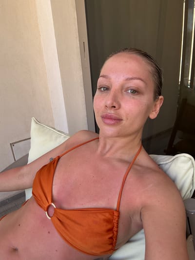 A light-skinned woman with wet blonde hair pulled back from her face is reclined in an outdoor setting. She is wearing an orange bikini top with a gold ring detail at the center. The location is inferred to be El Gouna, Egypt, although there are no specific identifiable landmarks in the frame. The background features a textured beige wall, a sliding glass door with a metal frame, and a cushioned outdoor chair. The overall composition is angled, giving a slight tilt to the scene. The woman's skin appears somewhat reddened, possibly due to sun exposure.