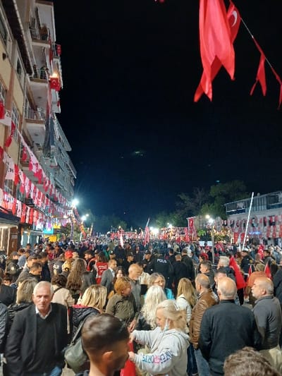 The image depicts a large crowd of people gathered at night in Silivri, Türkiye (latitude: 41.072, longitude: 28.244). The street is lined with buildings and decorated with Turkish flags. The crowd is dense, with people of all ages and genders, and many are holding red Turkish flags. Some people are wearing police uniforms. There are also multiple light poles along the street with street lights illuminating the scene. A large building can be seen in the background, with an awning and a light above the doorway. The crowd is mostly stationary and appears to be gathered for a celebration or event.