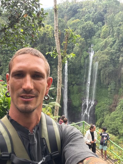A man poses for a selfie in front of a waterfall. He has short blond hair and large, decorative ear gauges. He wears a grey shirt with straps and a backpack. Behind him, lush green foliage covers the landscape with several streams of water flowing down. Other people stand on a viewing platform to the right of the frame, enjoying the waterfall views.