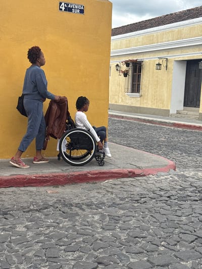 A person in a wheelchair is situated on a cobblestone street next to a yellow building with a street sign reading "4a. Avenida Sur". Another individual is helping the person in the wheelchair, handing them a brown jacket. The buildings feature traditional architectural elements with dark wooden doors and window boxes. The street appears slightly sloped, and the sky overhead is overcast.