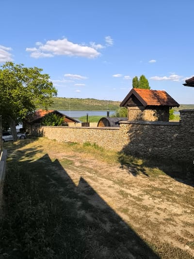 The image depicts a sunny outdoor scene in Jora de Mijloc, Moldova, featuring a rustic stone-walled compound next to a body of water, likely the Jora de Mijloc Reservoir. The compound contains two small buildings with reddish-brown tiled roofs, constructed with rough-hewn stones. One building is larger, resembling a small house or outbuilding, while the other is a smaller, more enclosed structure with a dark roof. A curved, barrel-shaped structure, possibly a sauna or storage, is visible behind the stone wall.

The stone wall itself is a prominent feature, built from irregularly shaped stones and stretching across the foreground and midground of the image. Beyond the wall, a wide, calm body of water is visible, bordered by lush green hills in the distance. The sky is a vibrant blue with scattered white cumulus clouds.

In the left of the image, a large, leafy green tree provides shade, and a silver car is parked near the building. The ground in the foreground consists of dry grass and dirt, with distinct shadows cast from an unseen structure or trees, creating a geometric pattern on the ground. The overall impression is one of a peaceful, rural setting, possibly a vacation or recreational area, with a focus on natural materials and integration with the landscape. There are no people visible in the image.