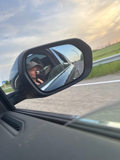The image shows a side mirror of a car reflecting a person wearing a cap and holding a phone. The background visible through the mirror suggests the car is moving on a road with green fields on the sides. The road is divided by a white line. The driver's reflection shows that they are in a car, and the rest of the scene seen in the mirror, including the sign, indicates the location may be in Fastovetskaya, Russia.