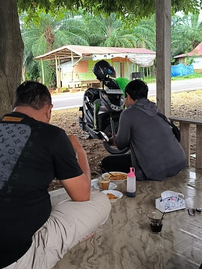In Teupinreusep, Indonesia, two men are seated on the ground under a covered area. One man, in the foreground and facing away from the camera, wears a black t-shirt with a grey patterned design and light-colored pants. He appears to be looking down at his food. The second man, also facing away from the camera, is seated to the right of the first man. He wears a grey hooded sweatshirt and dark pants, and is looking at a mobile phone he is holding. In front of them on a tiled surface, there are two plates of food, one with noodles, a plastic bottle with a pink lid, a box of tissues with a floral design, a glass of dark liquid with a spoon, and a pair of glasses. A motorcycle with a black helmet and a green bag is parked to the left of the second man. The background shows a rural landscape with palm trees, a small building with a tin roof, and a wooden fence. The overall impression is of a casual meal break in a tropical setting.