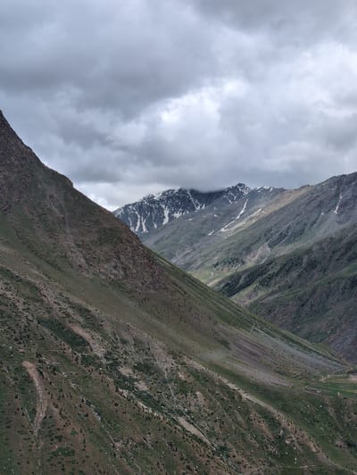 A vertical, medium shot captures a dramatic mountain landscape in Darcha, India, under a heavily overcast sky. The foreground is dominated by a steep, rocky mountainside covered in sparse green vegetation and scattered rocks. This slope extends diagonally from the lower left corner upwards and towards the right. In the mid-ground, a deep valley is visible, separating this foreground slope from a series of further mountain ranges. These distant mountains are also steep and rugged, with patches of snow clinging to their higher elevations. A winding path is faintly discernible on one of the mid-ground slopes. The sky is filled with thick, dark gray clouds, obscuring any direct sunlight and casting a somber mood over the scene.
