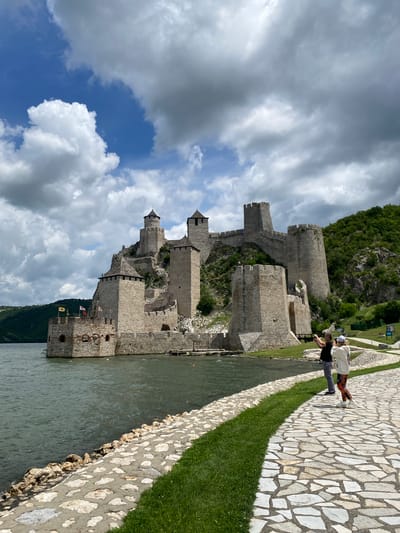 The image shows Golubac Fortress, a medieval castle located on the Danube River in the Golubac Municipality, Serbia. The fortress is situated on a rocky outcrop, with several towers and walls visible. The Danube River flows in front of the fortress, and the surrounding landscape is hilly and covered in vegetation. There are two people walking on a paved path in the foreground, one of whom is taking a photograph of the fortress. The sky is cloudy with some patches of blue.