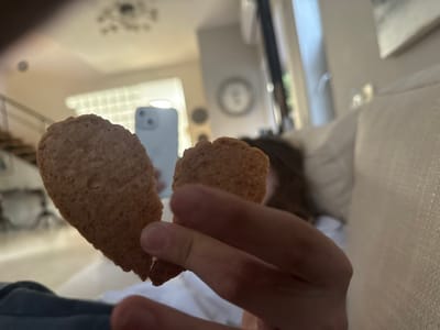 The scene is set in an indoor residential space, likely a living room, within the Menashe Regional Council, Israel. In the foreground, a light-skinned hand holds two light-brown, textured, elongated cookies. The cookie on the left is larger and appears to be in two pieces or partially broken at its narrow end. The cookie on the right is smaller and held between fingers. In the mid-ground, a person with dark hair is partially visible, resting against a white upholstered sofa on the right side of the frame. In the background, a light blue/grey smartphone is held upright, reflecting a bright light source. Further back on the left, a modern staircase with dark treads and light railings ascends. A wall with a grid-like pattern, possibly a window with a screen or blinds, is visible behind the staircase. A decorative chandelier is suspended from the ceiling in the upper center of the frame. The room is brightly illuminated.