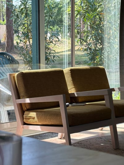 A two-seater sofa with olive green upholstery and light wood frame sits in front of a large window overlooking a green outdoor space with trees. A smartphone is resting on the right cushion of the sofa. Sunlight streams through the window, casting shadows on the sofa and the floor. A patterned rug is visible beneath the sofa. The overall setting suggests a residential interior in Heijen, Netherlands.