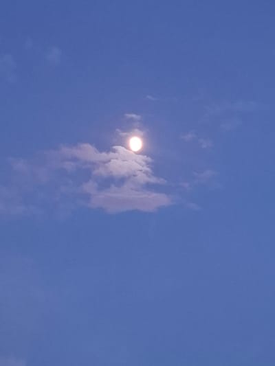 This media file depicts a bright, full moon in a clear, blue sky. The moon is a prominent subject, appearing as a luminous circle with a slight reddish hue. It is partially obscured by wispy, white clouds that are illuminated by the moon's glow. The clouds are scattered across the sky, creating a dynamic and ethereal atmosphere. The sky is a deep blue, suggesting it is either late evening or early morning. There are no people or man-made objects visible in the image. The overall scene is serene and tranquil, with the moon and clouds creating a beautiful celestial display. The location context is Urus-Martan, Russia.