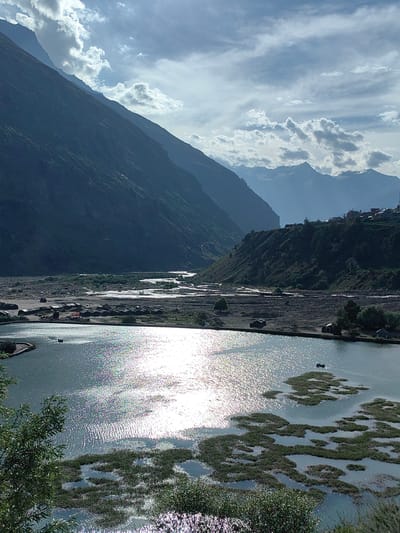 The image depicts a panoramic view of the Kullu Valley in Yamling, India, on a partly cloudy day. In the foreground, a calm body of water reflects the bright sunlight, creating a shimmering effect. Patches of green vegetation emerge from the water, forming islands. Two small boats are visible in the distance on the water.
Beyond the water, a wide, dry riverbed stretches into the distance, with a meandering river flowing through it. Along the banks of the riverbed, several small, dark structures, possibly tents or small huts, are scattered. Further in the background, a rugged, verdant mountain slopes steeply upwards, with a village nestled on its upper slopes, featuring light-colored buildings. In the far distance, a range of majestic, snow-capped mountains are silhouetted against the sky. The sky itself is a mixture of bright blue and dramatic, voluminous clouds. The overall scene conveys a sense of natural grandeur and remote tranquility.