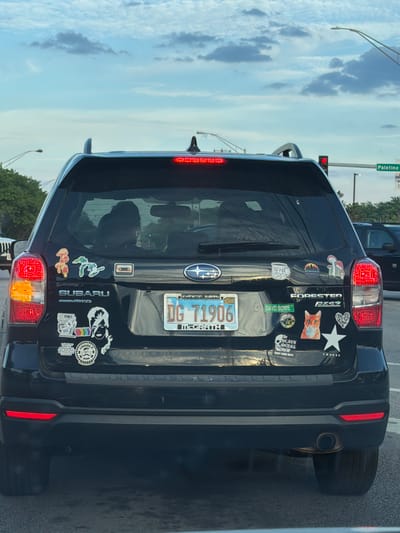 This image, rotated 90 degrees to the left, captures the rear of a black Subaru Forester SUV stopped at what appears to be a red traffic light, with other vehicles partially visible in traffic. The scene is set on a clear, daytime afternoon or early evening with scattered clouds in the sky.
The Subaru is extensively adorned with a variety of bumper stickers on its tailgate and rear window. Notable stickers include a green rectangle with "DAVID BOWIE," a sticker for "the HEAVY METAL SHOP SALT LAKE CITY," and "BYRNE'S." Other visible stickers depict a cat wearing glasses, a white star, a cassette tape, mushrooms, and various abstract or artistic designs, including one that appears to say "COPENHAGEN" on a coffee mug. The car's model "FORESTER" is prominently displayed, along with "SUBARU AWD." A custom exhaust pipe is visible on the lower right of the vehicle.
The Illinois license plate, "DG 71906," has a "02-27" registration tag and mentions "EVANSTON SUBARU" and "MCGRATH," likely indicating the dealership. A street sign in the background clearly reads "Palatine," providing a specific location cue for Arlington Heights, USA. The driver's and a passenger's reflections are faintly visible in the rear window.